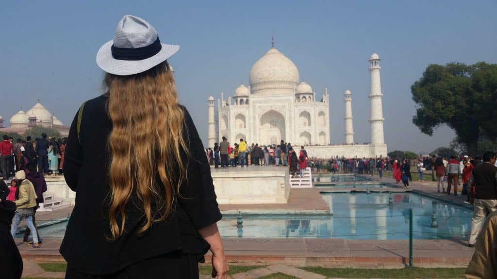 A woman wearing all black clothing with long wavy blond hair and a pale periwinkle fedora with a black band stands with her back to us, facing the Taj Mahal. 