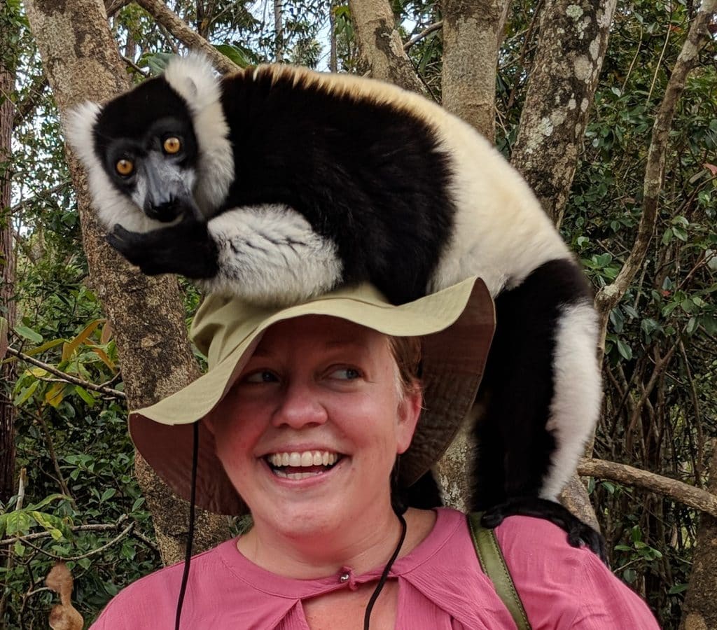 A woman in a pink blouse and tan safari hat smiles while a black and white lemur perches on her shoulder and head.