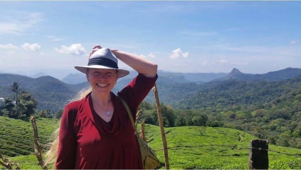 A woman in a maroon cowl-neck blouse stands outside in the sunlight with green rolling hills all around her and blue sky above. She holds her periwinkle straw fedora with a blank band on her head with her left hand.