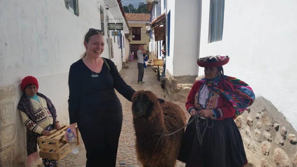A tall blond woman wearing all black smiles and places her left hand on a brown llama led by a native Peruvian woman in the streets of Cusco
