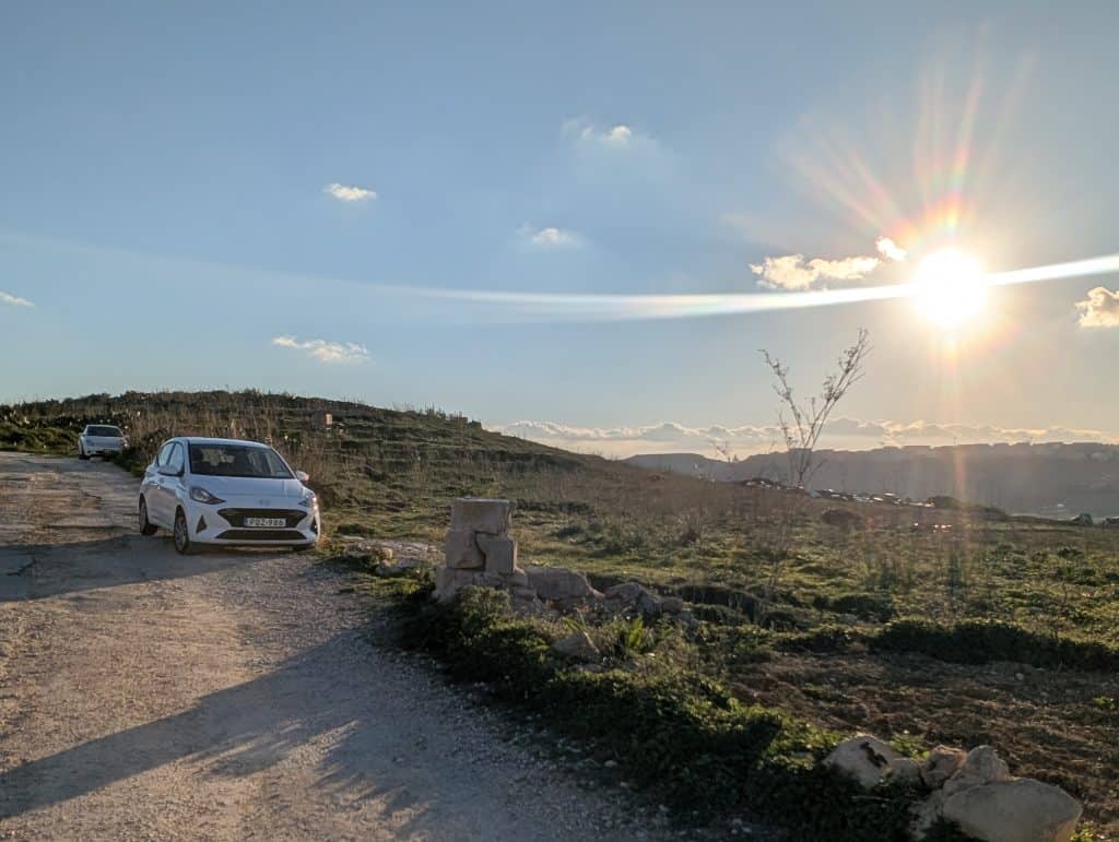 A small white compact car is parked on the side of a dirt road in Malta. Green hills and a blue sky unfurl around the vehicle with a bright sun at top right. 