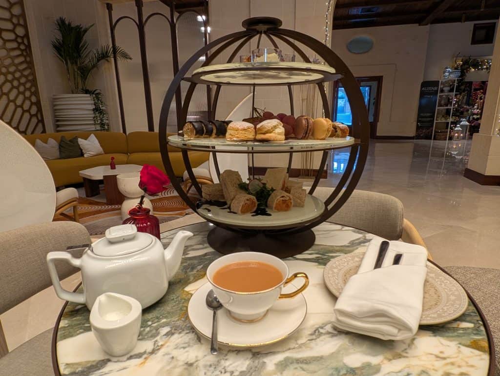 Afternoon tea at Kempinski San Lawrenz on Gozo. A spherical wrought iron cage with an open front and three tiers sits on top of a silver marble table. On the bottom tier are finger sandwiches. On the middle tier are scones, clotted cream, and jam. On the top tier is an array of small cakes and pastries. Around the tea tower are a white ceramic pot of tea, a small white ceramic pitcher of milk, and a a cup of black tea with milk stirred in resting atop a saucer with a tea spoon by its side. 