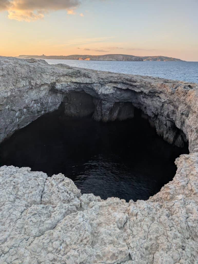 Coral Lagoon in the north of Malta at sunset—a dark hole in the white limestone cliffs reveals dark blue water with the luminous ocean stretching away offshore under a colorful sunset sky.