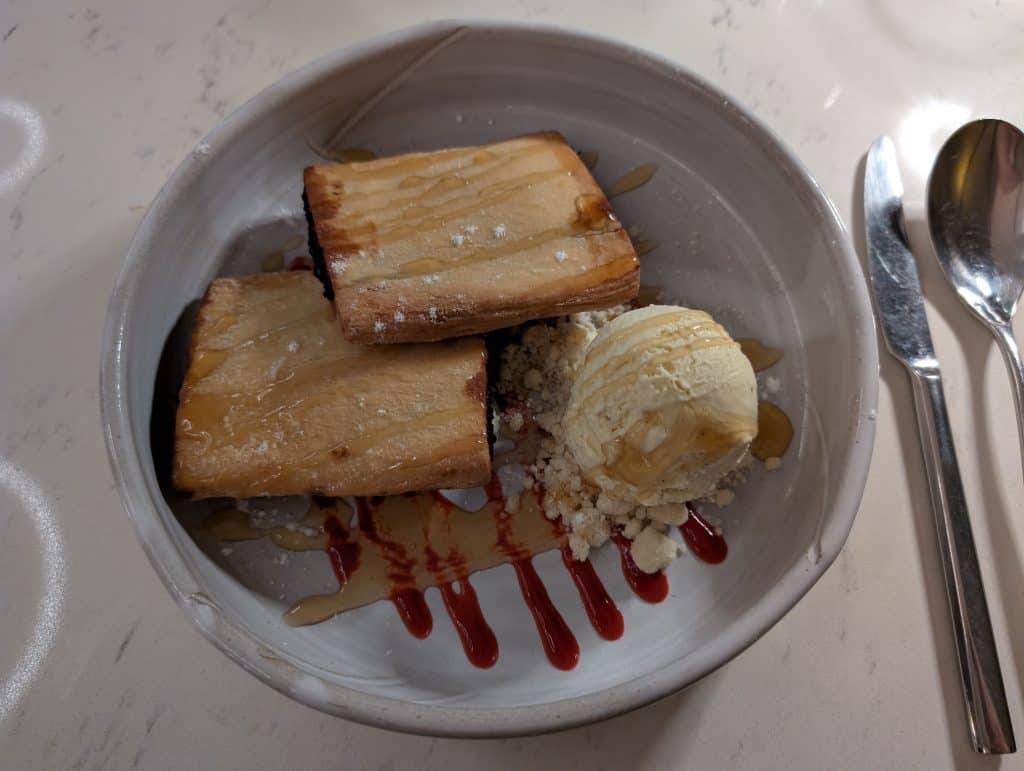 Two pieces of imqaret, a traditional Maltese desert, are stacked in a white bowl next to a scoop of vanilla ice cream and drizzled with honey and caramel. The desert is a flat rectangular pastry filled with sweet date paste. 
