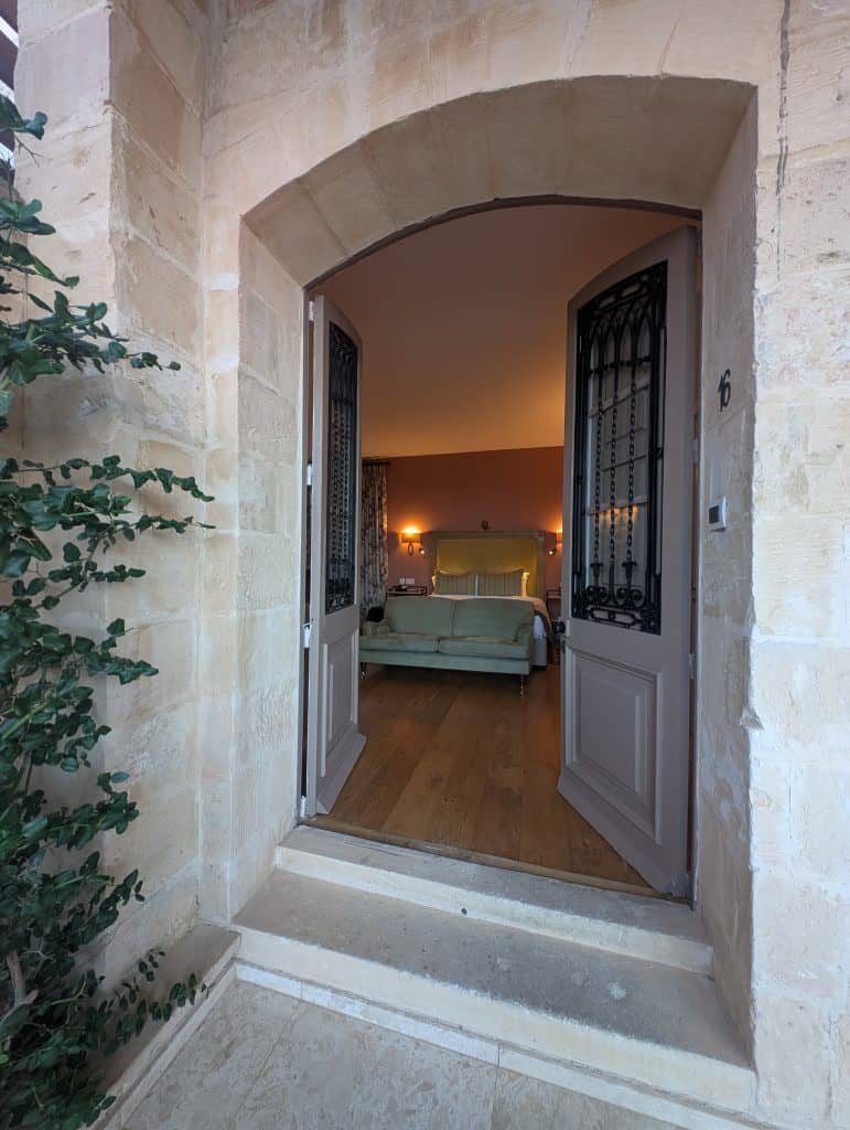 In a smooth white stone wall, double doors with wrought iron window bars open into a beautifully appointed luxury hotel room. There is a subdued seafoam green silk headboard, sconce lamps on either side of the bed, and a dusty mauve wall. 