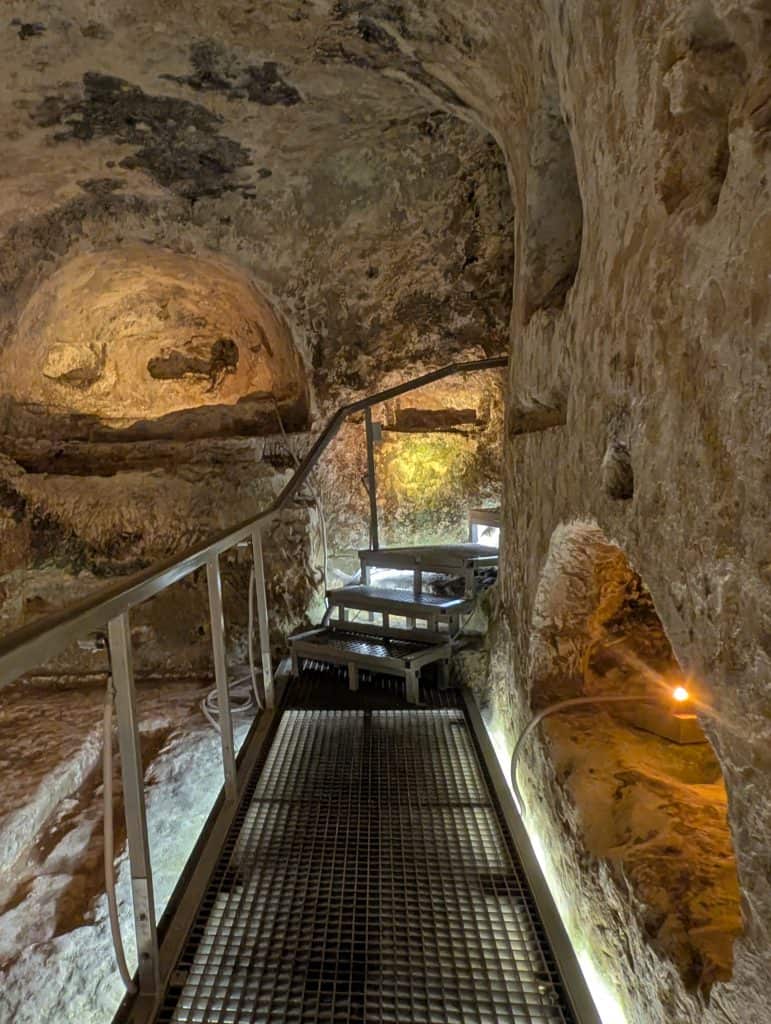 A steel walkway leads through an ancient stone catacomb complex with dark tunnels branching out on either side. The way is lit by lights hidden in the stone. 