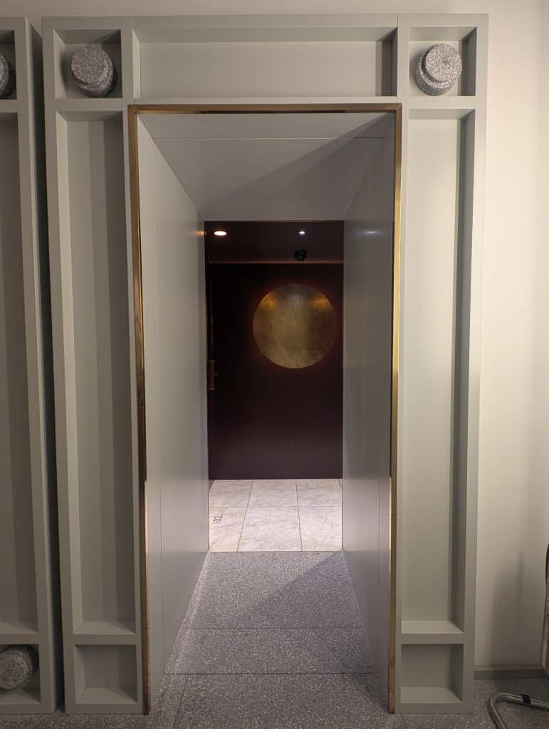 A hallway to rooms at Rosselli Hotel in Valletta, Malta. Grey molding frames an open portal down the hall to a burgundy wall with a large golden orb painted on it. The floors are silver marble. 