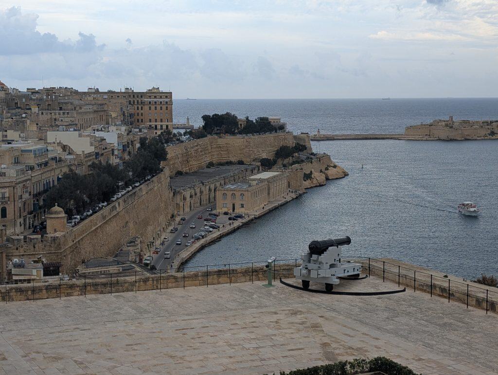 The view across the harbor from Upper Barakka Gardens in Valletta, Malta. The golden stone city unfurls to the left and beyond it is the open sea. In the foreground below is a black historic cannon. 