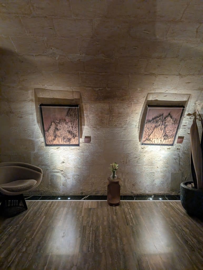 Essensi Spa at Iniala Harbor House. Ancient curved stone walls are illuminated by atmospheric lighting. Art hangs on the walls, a chair stands to the left, and a vase with flowers in it stands on the floor in the center of the wall. 