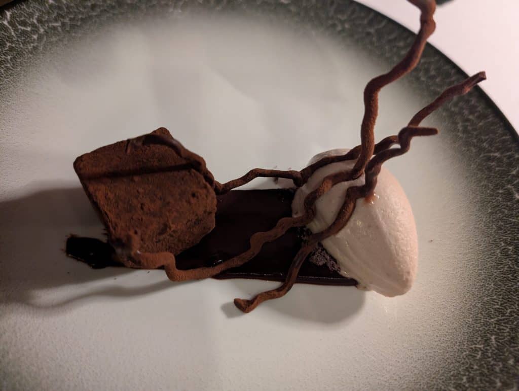A close-up image of a small, square chocolate brownie next to a scoop of olive ice cream and topped with artful chocolate sticks that resemble tree branches. 
