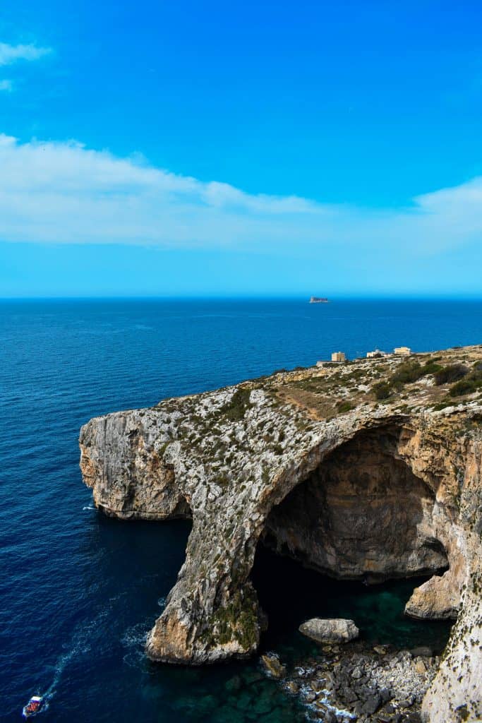 The Blue Grotto in Malta—a network of white stone sea caves extends inward under a stone arch. The arches rise out of calm blue water with a blue sky above. 