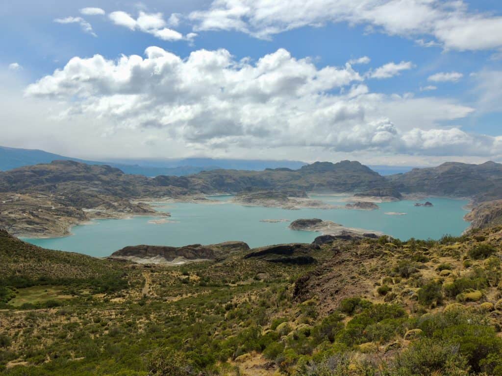 A teal glacial lake dotted with rocky silver outcroppings sprawls under a blue sky full of fluffy white clouds. A hilly green landscape surrounds the water in Carretera Austral, Chile. 