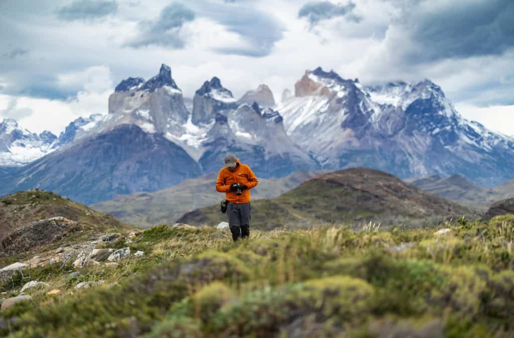A hiker in a neon orange jacket walks toward the photographer while looking down at his camera. Behind him, the jagged silver peaks of the Torres del Paine Massif in Chilean Patagonia tower up, covered in snow. A cloudy sky is above and hillocks of green grass are below. 
