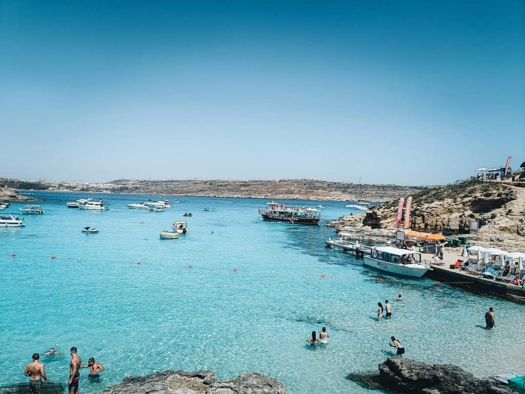 The Blue Lagoon on Comino Island, Malta. Bright powder blue water surrounds a white, rocky cove. A small boat sits at the dock and swimmers lounge in the clear, shallow water.
