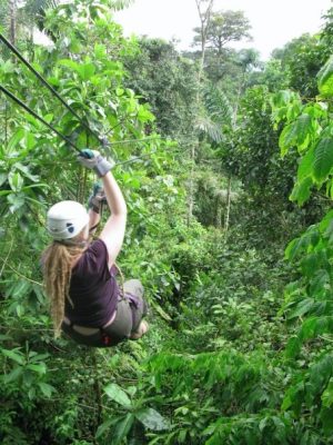 A blond woman with dreadlocks wears cargo pants, a purple T-shirt, and a white helmet. She faces away from the camera, suspended on a zip line that leads away into the jungle.